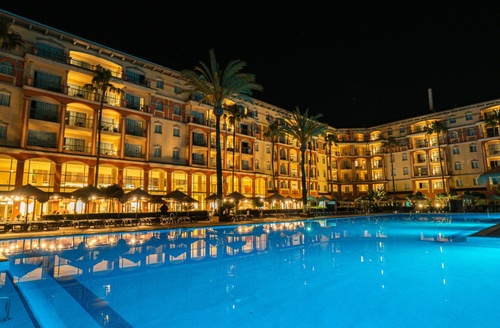 Hôtel tout compris à Islantilla : piscine illuminée et bâtiment avec palmiers la nuit.