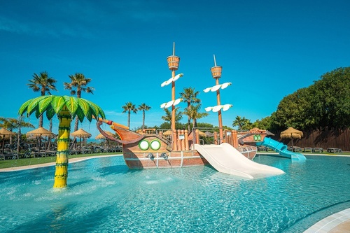 A vibrant water park scene shows a pirate ship play structure with slides and a palm tree water sprinkler in a pool, surrounded by sun loungers and palm trees under a clear blue sky.