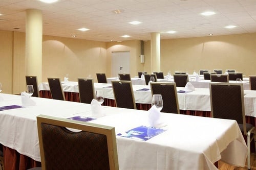 A brightly lit conference or meeting room is meticulously arranged with rows of white-clothed tables, chairs, and individual place settings including wine glasses and pamphlets.