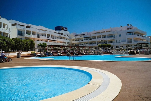 A resort with multiple white buildings, several swimming pools, and lounge chairs with umbrellas is visible under a clear blue sky.