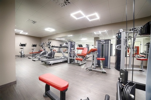 This image shows a modern gym equipped with various exercise machines, including treadmills, stationary bikes, and weight equipment, with large mirrors reflecting the interior along one wall.