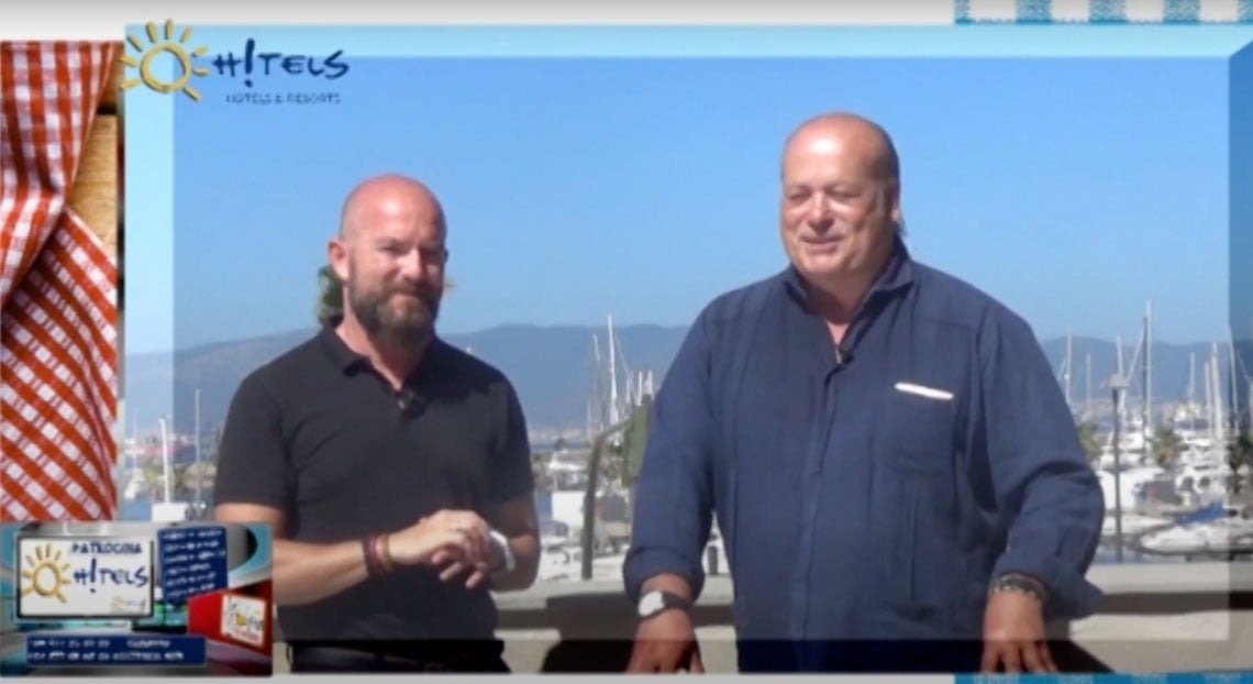 La imagen muestra la moderna entrada de cristal de un edificio con el letrero "GRAN HOTEL ALMERIA" en su fachada de azulejos grises, flanqueada por plantas en macetas.