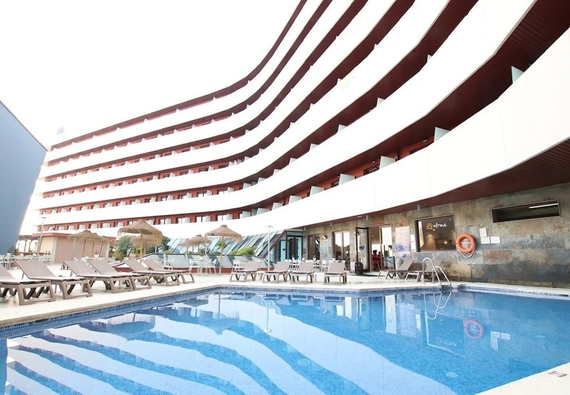 A imagem exibe a fachada moderna da entrada do Gran Hotel Almeria, com paredes de azulejos cinza claro, portas de vidro e vasos com plantas decorativas.