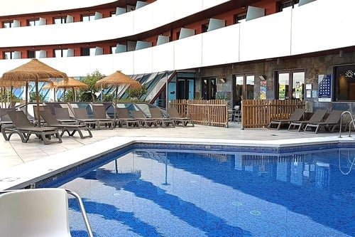 Una escena de piscina con tumbonas y sombrillas de paja frente a un edificio de hotel moderno con balcones curvos.