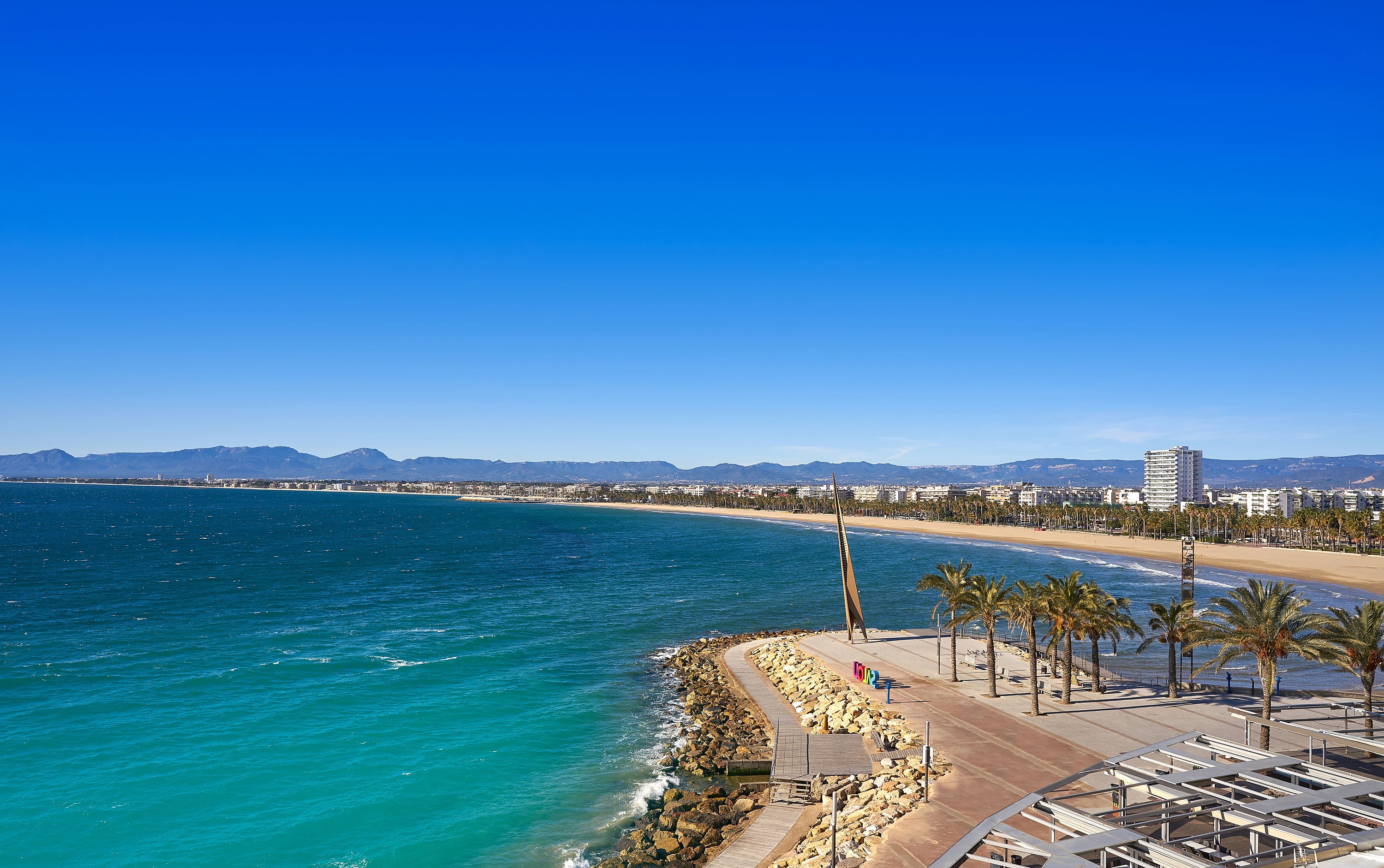 Coastal view from hotel: sandy beach, turquoise sea, palm trees, and city skyline. Ohtels: Corporativa