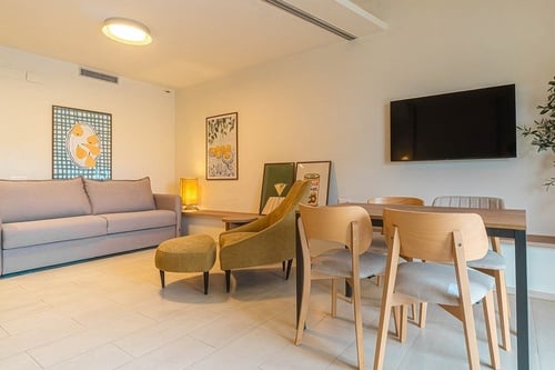 Un espacio combinado de salón y comedor con un sofá gris, un sillón mostaza, un televisor en la pared y una mesa con sillas de madera.