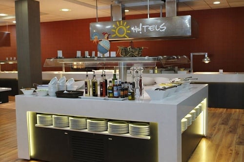 A brightly lit hotel buffet counter displays various condiments, white serving bowls, and stacked plates, topped by an overhead "Hi! HOTELS" sign and a decorative rooster, all set against a warm red wall and wooden floor.