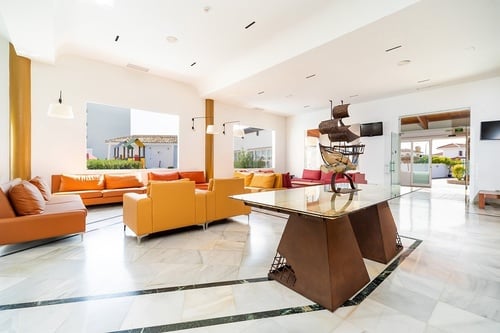 Un luminoso y moderno vestíbulo de hotel o sala de espera presenta sofás naranjas, amarillos y rojos, un suelo de mármol con detalles oscuros, grandes ventanas que dan a un patio con parque infantil y una mesa central con una maqueta de barco.