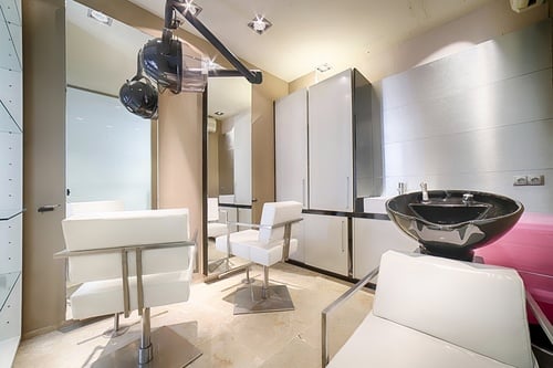 Este es un moderno salón de peluquería con asientos blancos, espejos, un lavacabezas y un secador de pelo colgante.