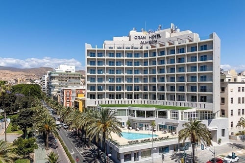 Le Gran Hotel Almeria, un grand bâtiment moderne avec de nombreux balcons et une piscine extérieure, borde une avenue plantée de palmiers, avec des montagnes en arrière-plan sous un ciel bleu.