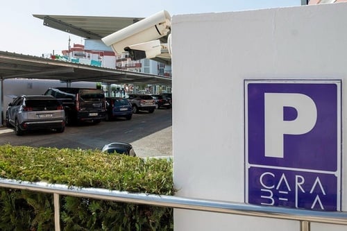 Aparcamiento exterior con coches bajo una estructura techada, cámaras de seguridad, un cartel de parking violeta con "CARABELA" y arbustos en primer plano.