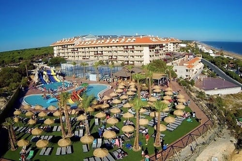 An aerial view captures a vibrant resort with a large hotel building, a lively water park featuring pools and slides, numerous sun loungers under straw umbrellas, and palm trees, all set against a backdrop of a dense forest and a long sandy beach by the ocean under a clear blue sky.