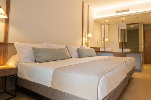 Una moderna y luminosa habitación de hotel cuenta con una cama grande, lámparas de lectura en la pared y un área de baño integrada con dos lavabos y espejos.