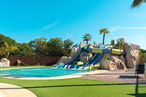 A vibrant water park features a swimming pool with multiple water slides, artificial rocks, palm trees, and artificial grass under a clear blue sky.