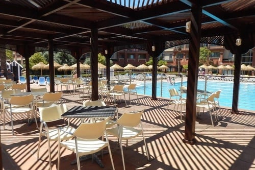 A wooden pergola covers an outdoor seating area with tables and chairs next to a swimming pool, casting prominent striped shadows on the ground, with resort buildings and thatched umbrellas in the background.