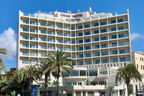 L'image présente un grand hôtel moderne, le "Gran Hotel Almeria", avec de nombreux balcons en verre et des palmiers devant, sous un ciel bleu clair.