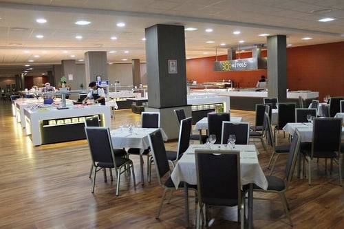 This image shows a modern, brightly lit buffet-style restaurant or dining hall featuring multiple food stations, set tables, and staff members.