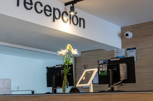 La imagen muestra una moderna recepción de hotel con el cartel "recepción" en la pared, equipada con ordenadores, una tableta y un florero con lirios blancos, bajo la vigilancia de una cámara de seguridad.