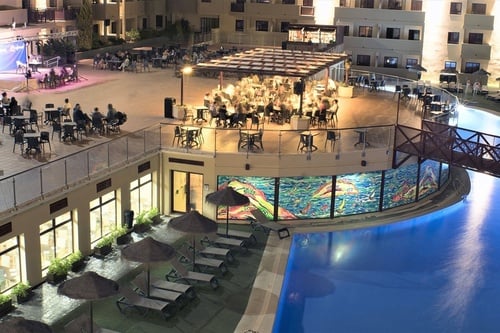 An illuminated hotel or resort complex is bustling with activity in the evening, featuring a tiered layout with a vibrant pool area, an outdoor dining and entertainment patio, and buildings in the background.