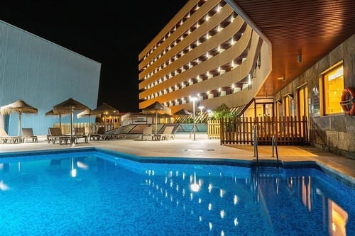 Vista nocturna de un complejo hotelero con una piscina azul, rodeada de tumbonas y sombrillas, y un edificio moderno con balcones iluminados.