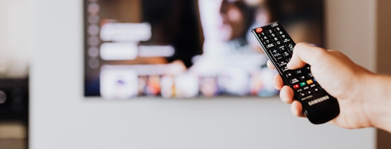 Une main tient une télécommande devant un écran de télévision flou.