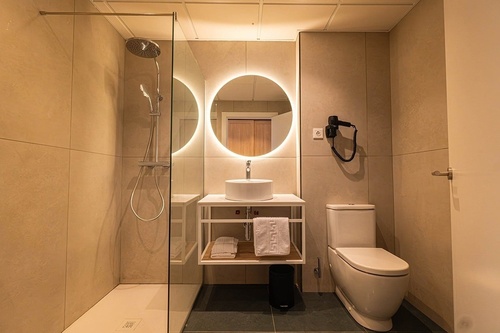 Este es un moderno cuarto de baño con una ducha de cristal, un lavabo sobre encimera con un espejo redondo retroiluminado y un inodoro, todo en tonos neutros.