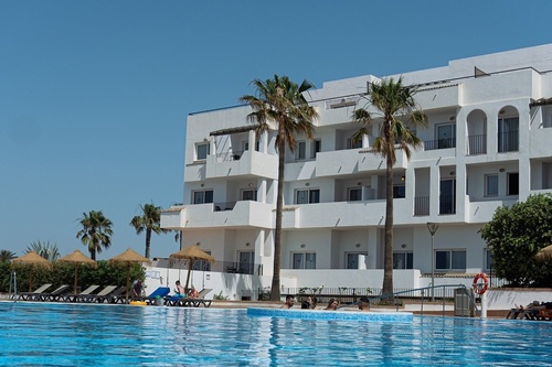 A white multi-story building with balconies and palm trees stands beside a blue swimming pool where people are relaxing, under a clear sky.