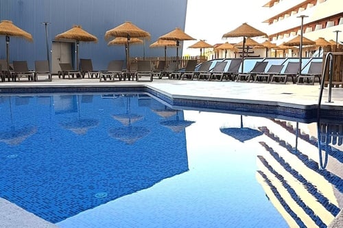 Una piscina azul rodeada de sombrillas de paja y tumbonas, con los reflejos de los edificios claramente visibles en el agua.