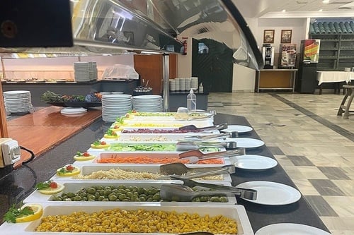 Un bufet de autoservicio presenta una variedad de ensaladas e ingredientes frescos en recipientes blancos con pinzas para servir, junto a pilas de platos limpios.