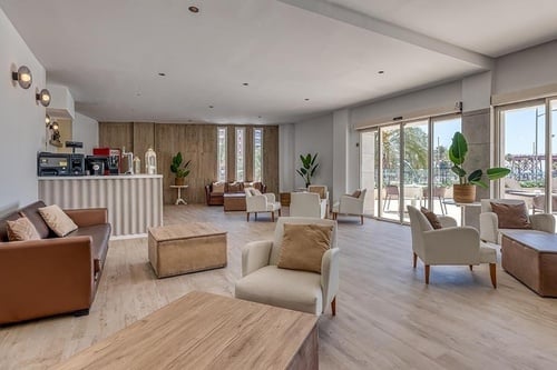 Un salon moderne et lumineux, aménagé avec un bar, des canapés et fauteuils confortables aux tons neutres, s'ouvre sur l'extérieur par de grandes baies vitrées.