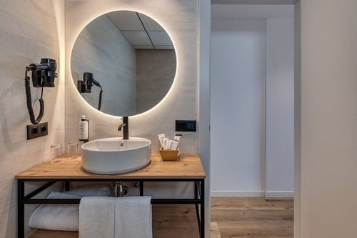 Une salle de bain moderne présente un meuble-lavabo en bois clair avec une vasque blanche, un miroir rond rétroéclairé et un sèche-cheveux mural, adjacente à un passage lumineux.