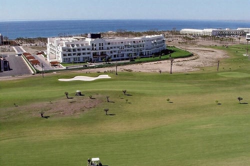 A sprawling golf course with several trees and golf carts lies in the foreground, with a large white building and other structures in the middle ground, and the vast blue sea stretching to the horizon under a clear sky in the background.