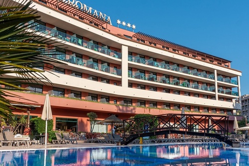 Un grand hôtel avec de nombreux balcons et l'enseigne "ROMANA" est visible au-dessus d'une piscine avec un pont en bois, sous un ciel bleu clair.