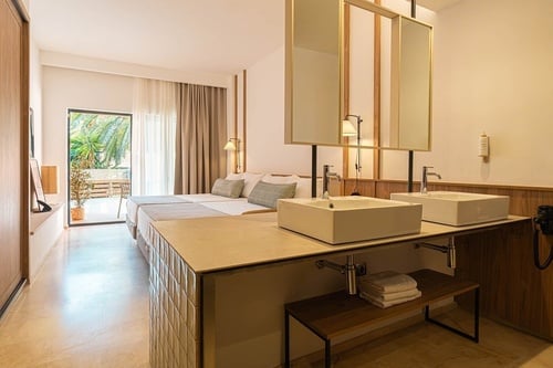 Una moderna habitación de hotel presenta dos camas, un balcón con vistas a la vegetación y un tocador doble con lavabos y espejos.