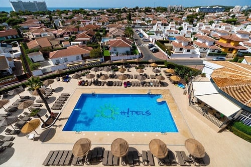 Una vista aérea muestra una piscina con el logo "Hi! Hotels" rodeada de tumbonas y sombrillas, con un barrio residencial y el mar al fondo.