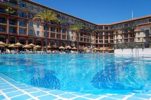 A low-angle shot captures the glistening blue water of a swimming pool in the foreground, with a large resort hotel, palm trees, and rows of straw umbrellas and sun loungers visible under a clear blue sky in the background.