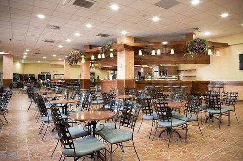 Un amplio comedor de restaurante o cafetería, con numerosas mesas y sillas metálicas vacías, un mostrador con taburetes al fondo y un suelo de baldosas con un patrón geométrico.