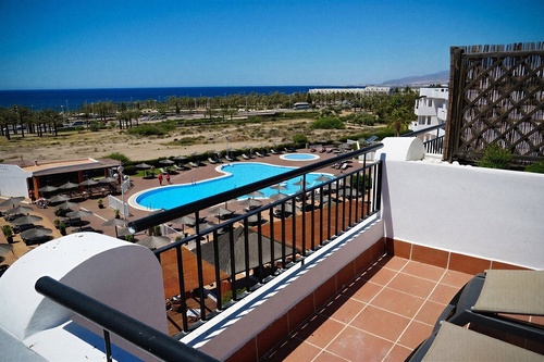 From a high-angle balcony, an outdoor resort swimming pool with sun loungers and palm trees is visible, stretching towards a clear blue ocean under a bright sky.