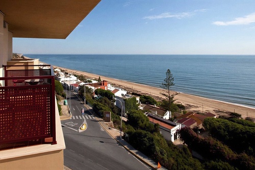 A high-angle view from a balcony reveals a scenic coastal landscape featuring a sandy beach, the calm ocean, a winding road, and houses nestled among green vegetation.