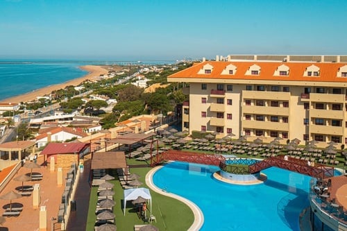 An elevated view captures a hotel and its extensive pool area, complete with sun loungers and parasols, set against the backdrop of a sandy beach, the ocean, and a coastal town.