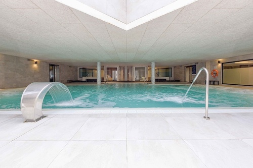 Une piscine intérieure lumineuse est dotée d'un plafond texturé, d'un carrelage clair et de deux jeux d'eau animant sa surface.