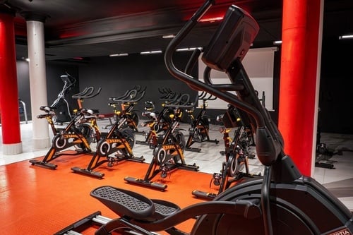 Se muestra una sala de fitness con varias bicicletas de spinning y una elíptica en primer plano, destacando sus pilares rojos y el suelo anaranjado.