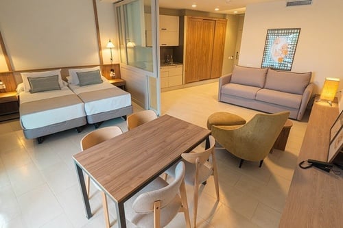 Un moderno apartamento tipo estudio integra un dormitorio con dos camas, una sala de estar con sofá y sillón, un comedor y una cocineta visible a través de una partición de vidrio.