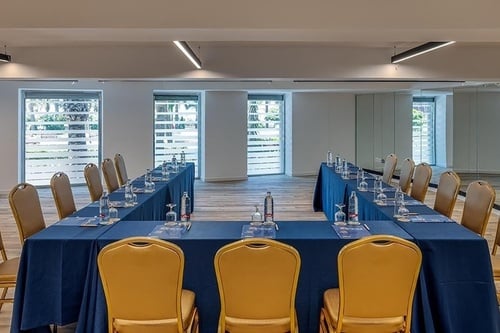Une salle de conférence moderne est aménagée en U avec des tables bleues et des chaises dorées, éclairée par des fenêtres donnant sur l'extérieur.