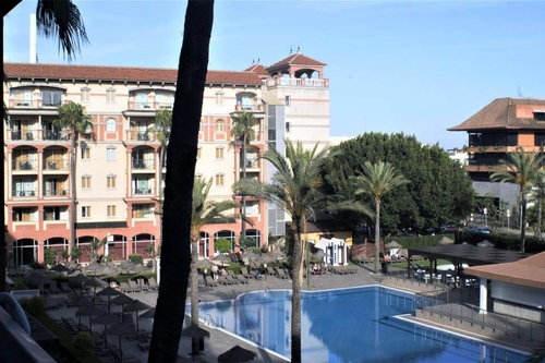 A large resort building with balconies overlooks a spacious swimming pool surrounded by palm trees and lounge chairs under a clear sky.