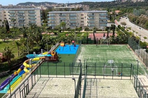 Una vista aérea muestra un complejo residencial con diversas instalaciones recreativas al aire libre, incluyendo toboganes acuáticos, varias canchas deportivas y un parque infantil, rodeado de zonas verdes.