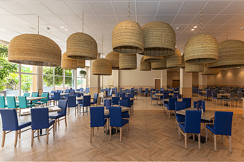 Espaço gastronômico de hotel com decoração moderna, mesas e cadeiras azuis, luminárias de vime e vista para jardim. Hotel tudo incluído em La Pineda | Ohtels La Hacienda