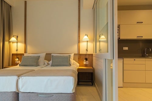 Una moderna habitación de hotel con dos camas individuales y una cocina americana visible a través de una partición de vidrio.