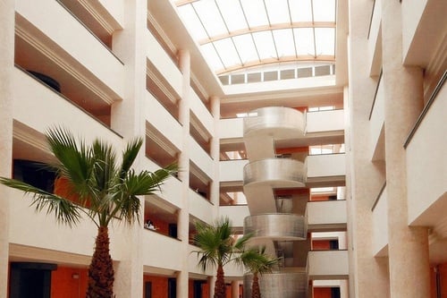An expansive, multi-story atrium is brightly lit by a skylight, showcasing tiered balconies, indoor palm trees, and a central spiral structure.