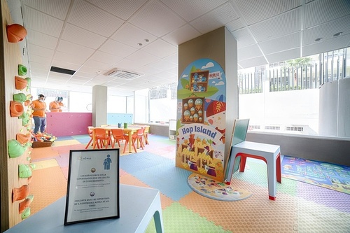 A brightly lit and colorful children's play area features a climbing wall, small tables and chairs, a "Hop Island" themed pillar, and a sign reminding of adult supervision.